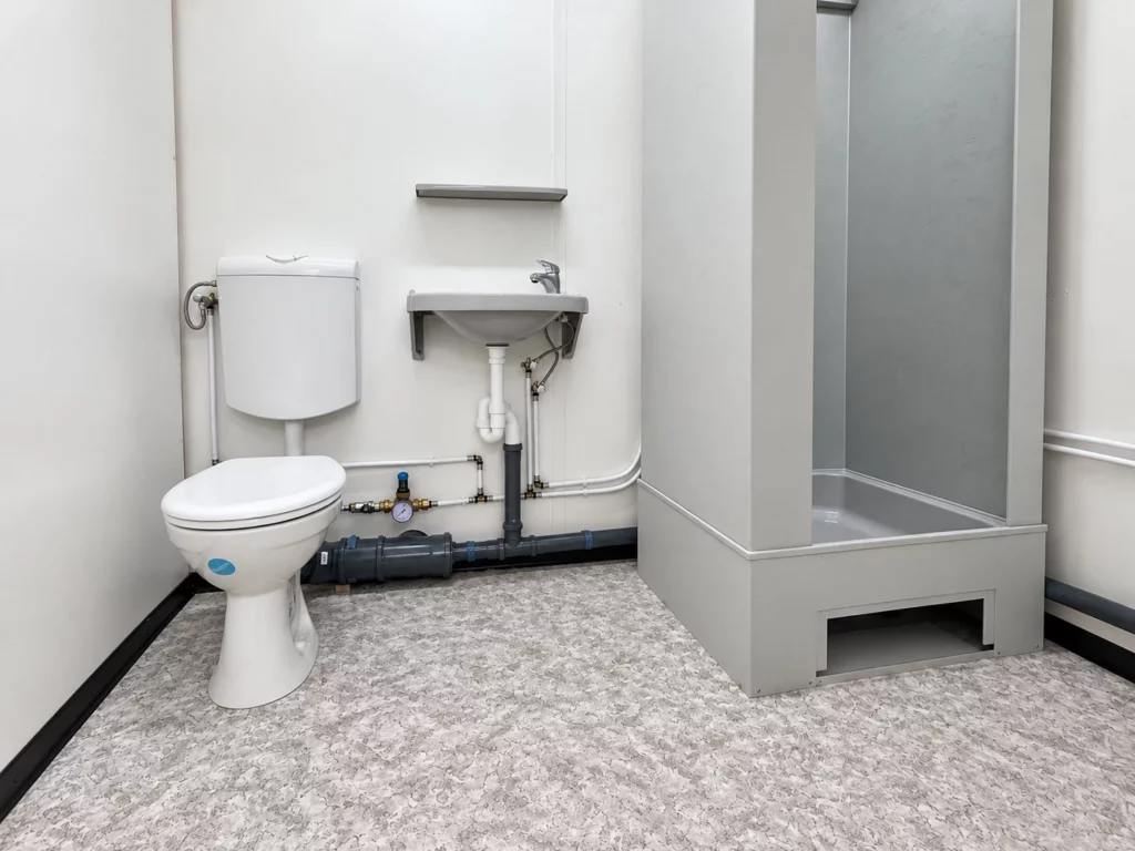 Sanitärraum mit WC, Waschbecken und Duschkabine, die Leitungen sichtbar an der Wand